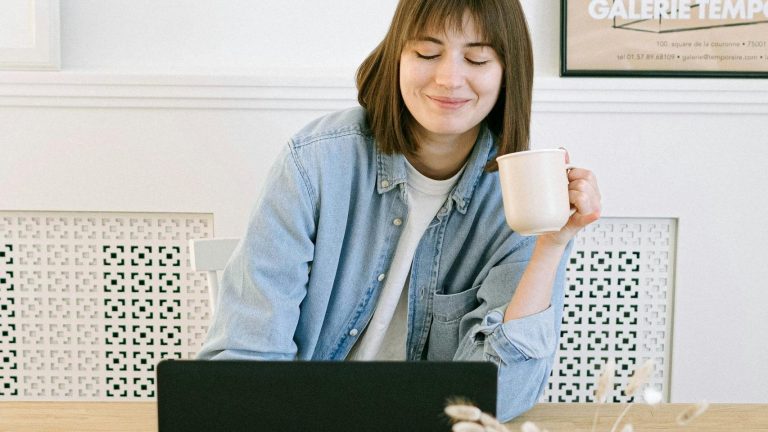 Woman in a home office setting, smiling with a coffee cup near a laptop.