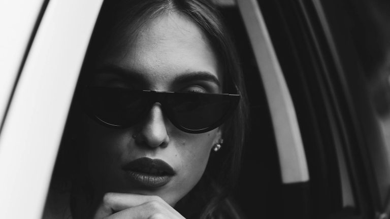 Chic portrait of a woman wearing sunglasses gazing out of a car window in black and white.
