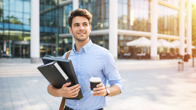 Young professional entrepreneur walking outside a modern office building holding business files and coffee in a bright morning setting.