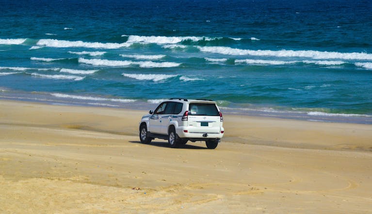 A white SUV traverses a picturesque sandy beach by the ocean under clear skies.