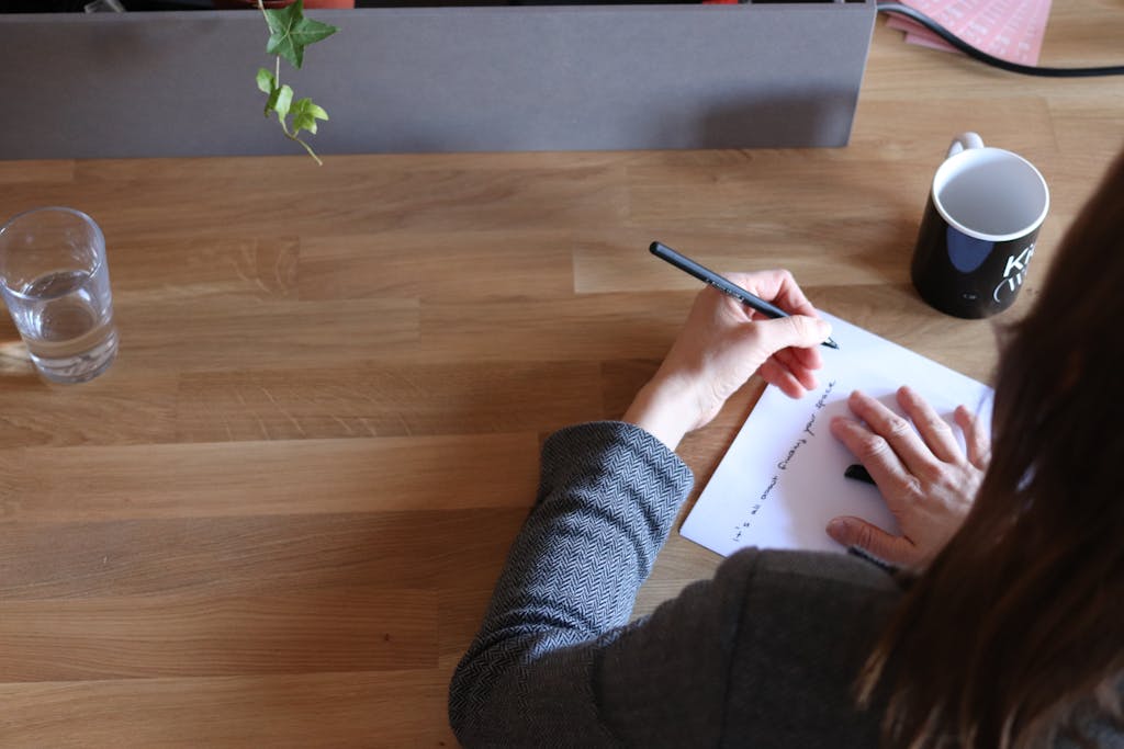 An adult woman taking notes on paper at a wooden desk in an office setting with a mug and glass of water.