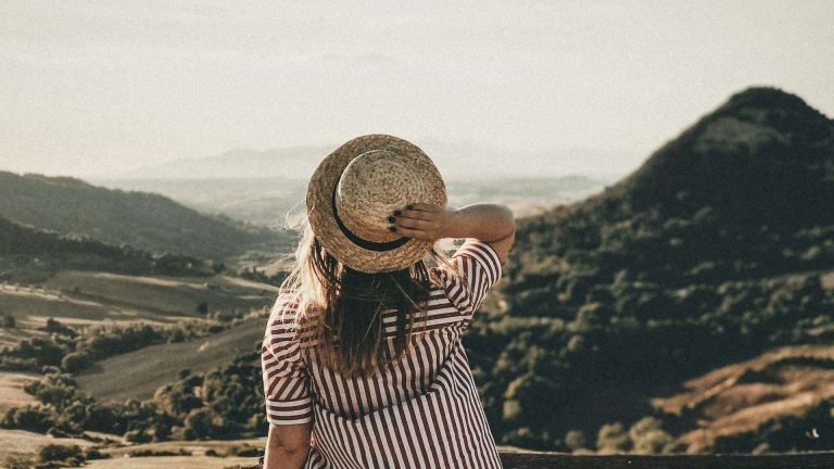 Young woman in striped dress and hat embracing a scenic countryside landscape during daylight.